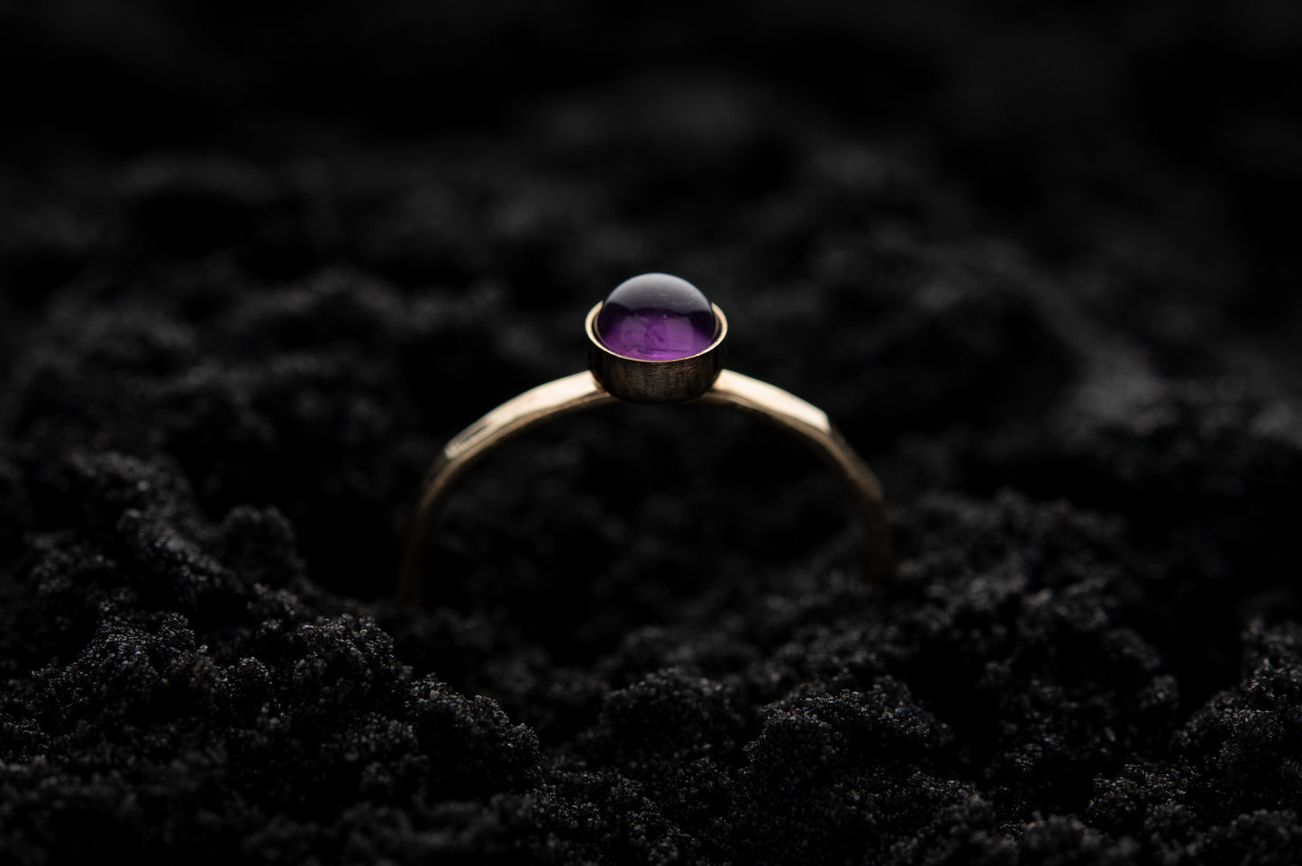 A single amethyst stacking ring with yellow gold band and around amethyst stone, rests gently in soft black sand..