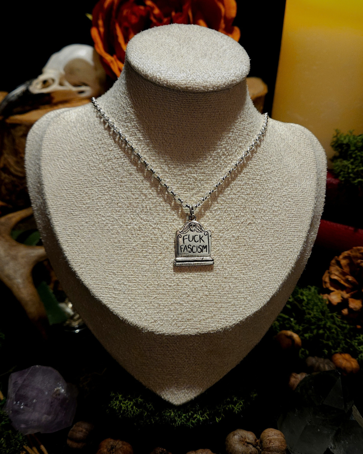 A .925 silver tombstone pendant featuring a skull, and engraved with 'FUCK FASCISM', is displayed on a beige vintage style bust. The display is set against a dark, mystical background with candles, a skull, crystals and other macabre and dark academia themed items