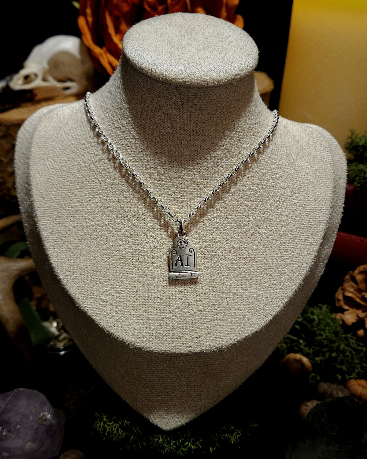 A .925 silver tombstone pendant featuring a skull, and engraved with 'FUCK AI', is displayed on a beige vintage style jewellery bust. The display is set against a dark, mystical background with candles, a skull, moss, crystals and other macabre and dark academia themed items