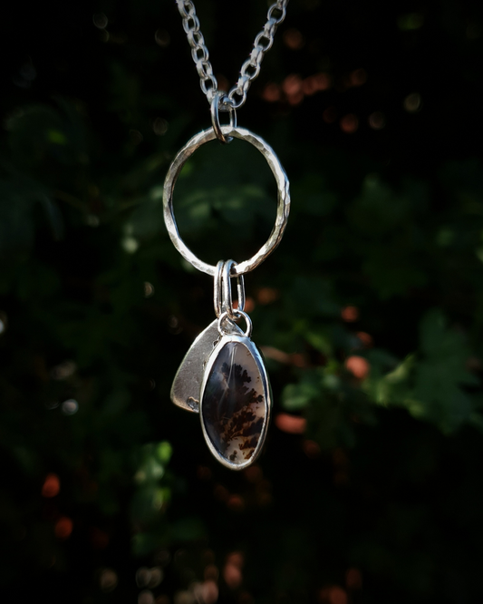 Silver pendant comprised of a silver circlet from which a solid silver triangle, imprinted with a cypress sprig, and dendritic agate stone pendant hang from a silver chain. The dendritic agate is translucent with dark brown dendrites. The background is out of focus natural greenery.