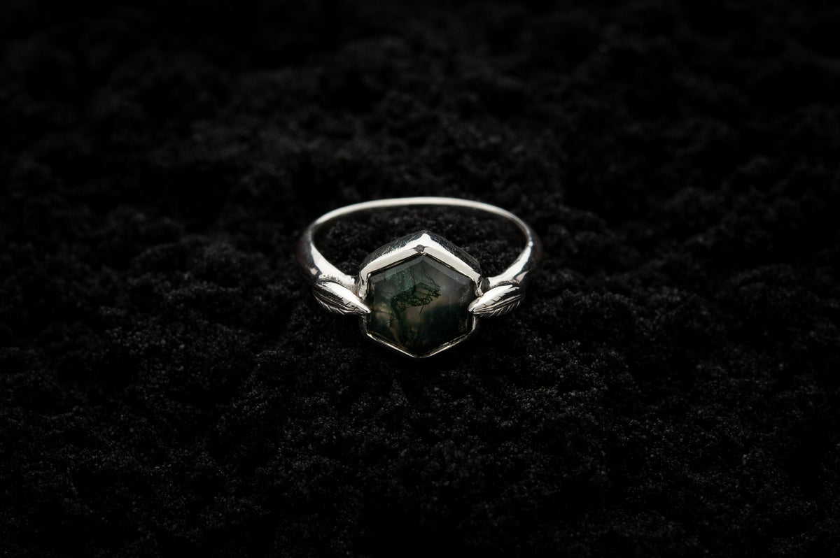 A leafy engagement ring rests on black sand and is lit in soft, glowy light.
A luscious hexagonal moss agate is set in a white gold setting with a slim leaf on either side, folded slightly over the stone.