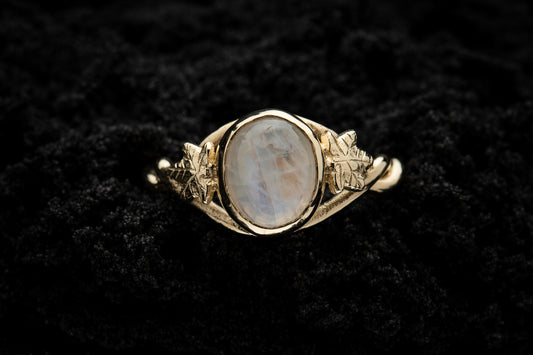 A botanical engagement ring made from rainbow moonstone and gold sits in black sand. It is photographed from above and features a twisted band and an ivy leaf on each shoulder. The moonstone shines gently in the camera flash, teasing it's bluey rainbow hues.