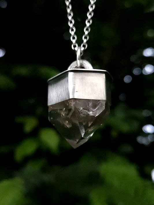 A chunky sterling silver pendant with a faceted garden quartz point, hanging on a belcher chain. Photographed outdoors against a blurred green foliage background. The image is taken from below to show the facets and inclusions in the quartz. The silver has a slightly matte, brushed finish.