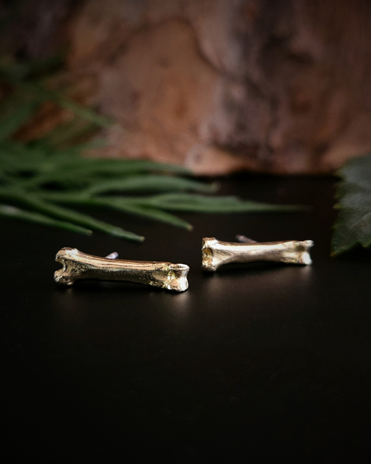 A pair of luxury gothic and witchy 9 carat gold bone stud earrings laying on their side on a black surface with a natural background of rough bark and green foliage.