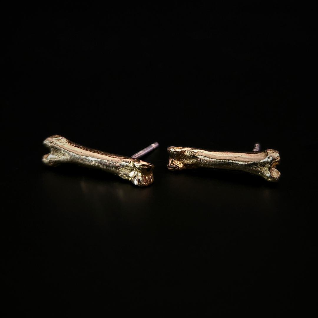 A pair of witchy 9 carat gold bone stud earrings laying on their side on a solid black background.