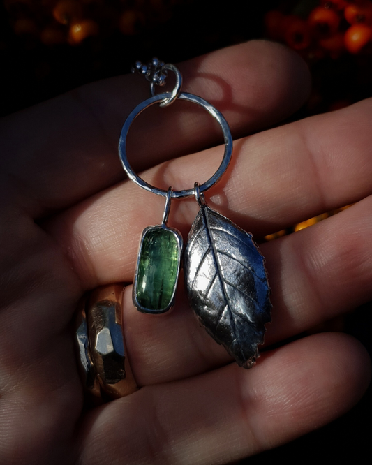 Silver necklace with a solid silver leaf and mint green stone pendant attached to a silver circlet. It is sitting in an open hand, which wears two gold rings, to demonstrate its size. There are red berries out of focus in the background.