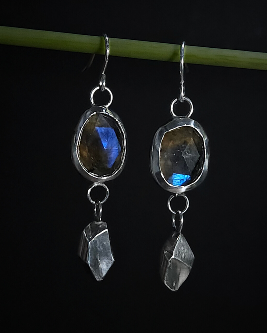 A pair of sterling silver and rose-cut faceted dangly earrings hang from a green plant stem. Their geometric and rough-cut drops underneath the gemstone are inspired by the slate mines and mountains of Wales.