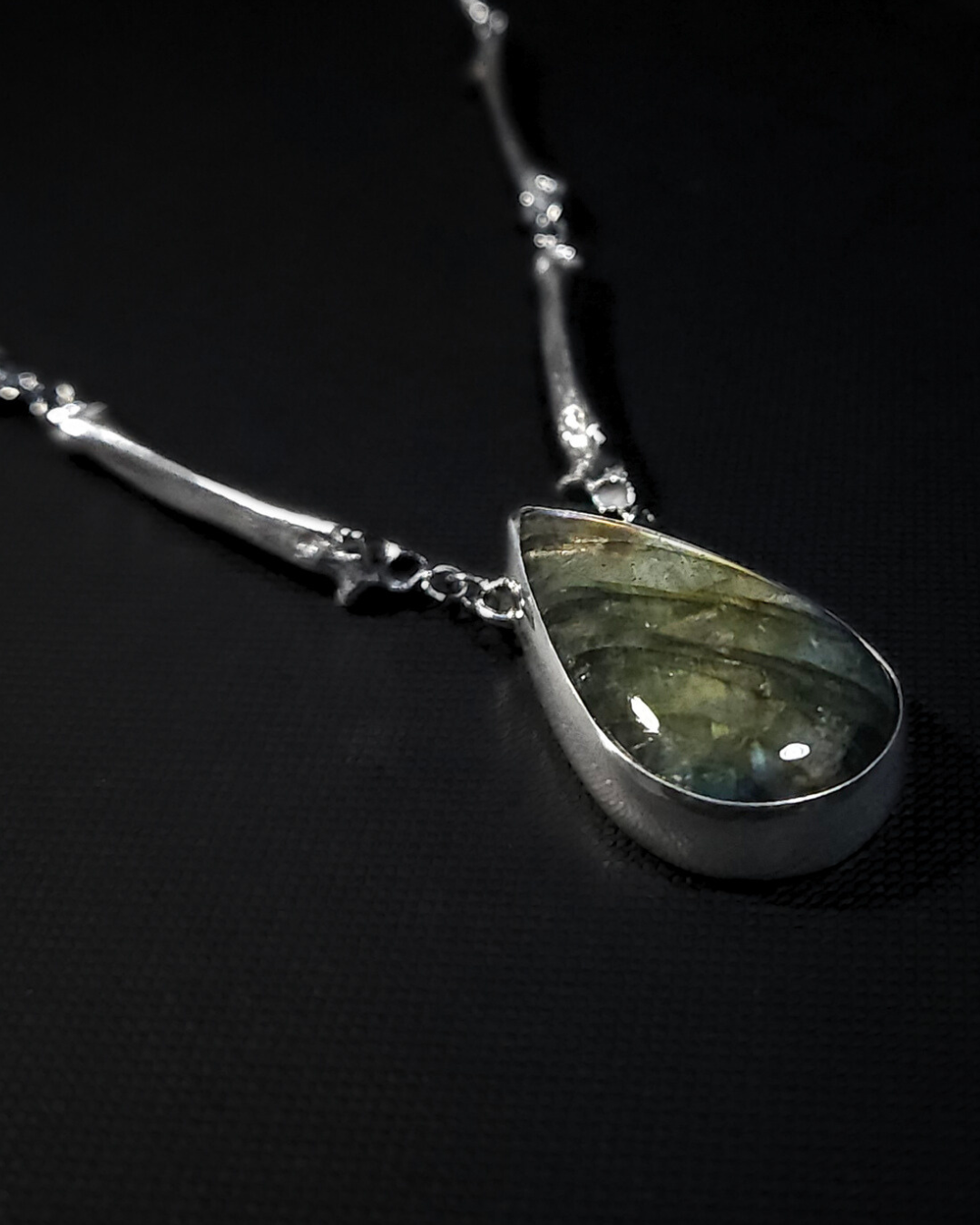 Close up shot of necklace with a teardrop-shaped labradorite pendant on a black background. The chain is made of solid silver bone castings.