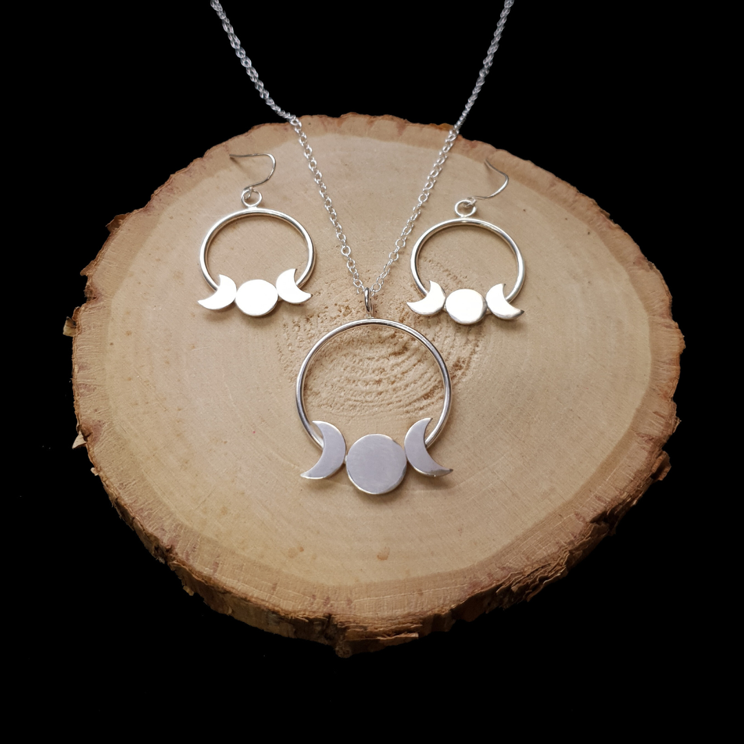 A set of sterling silver jewellery including dangly earrings & a necklace displayed on a wooden surface. The silver is polished to a high shine. Each pieces features the triple moon goddess on a large loop.