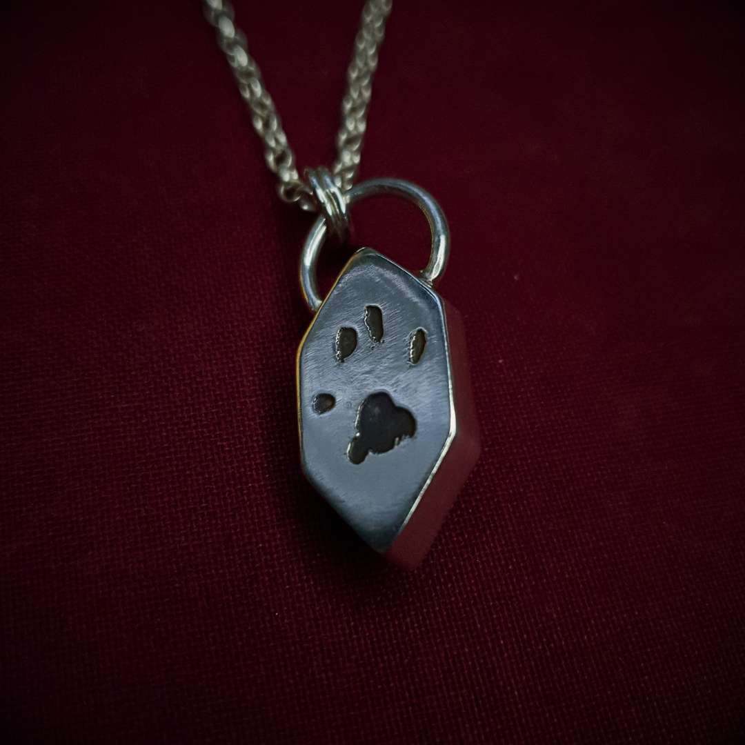 The reverse of a Memento Mori pendant is visible. It is a long hexagonal design in sterling silver. A pawprint has been relief-engraved in the back and oxidised black to show the finer details. The necklace is displayed against a dark red background.