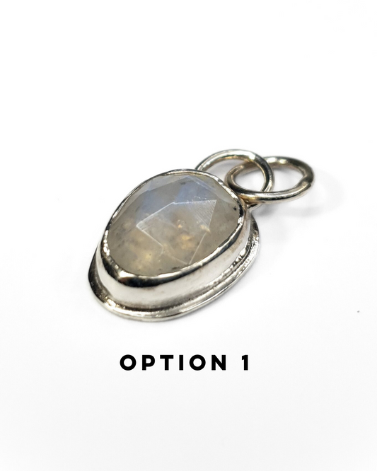 An irregular, oval-shaped rainbow moonstone is bezel set in silver and sits on a white background. The gemstone is rose-cut and the facets shine in the light displaying a beautiful show of rainbow iridescence. There is a large circular silver bail at the top with a similarly large jump ring attached. There is text below which reads "OPTION 1"