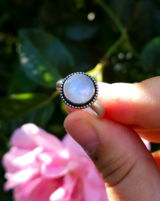 A sterling silver and round rainbow moonstone held by a hand against a blurred natural background. An etherial, blue, luminescence is visible in the high quality, ethically sourced stone.