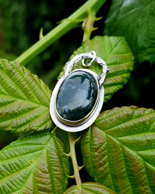 A rich, green, translucent moss agate sits in an oxidised silver bezel. The bail loop of the pendant is a highly textured casting of a twig in solid silver. The pendant is lightly speckled in rain drops and photographed gently balancing on the bed of bramble leaves in a blackberry bush. The whole image is luscious green and bathed in natural light.