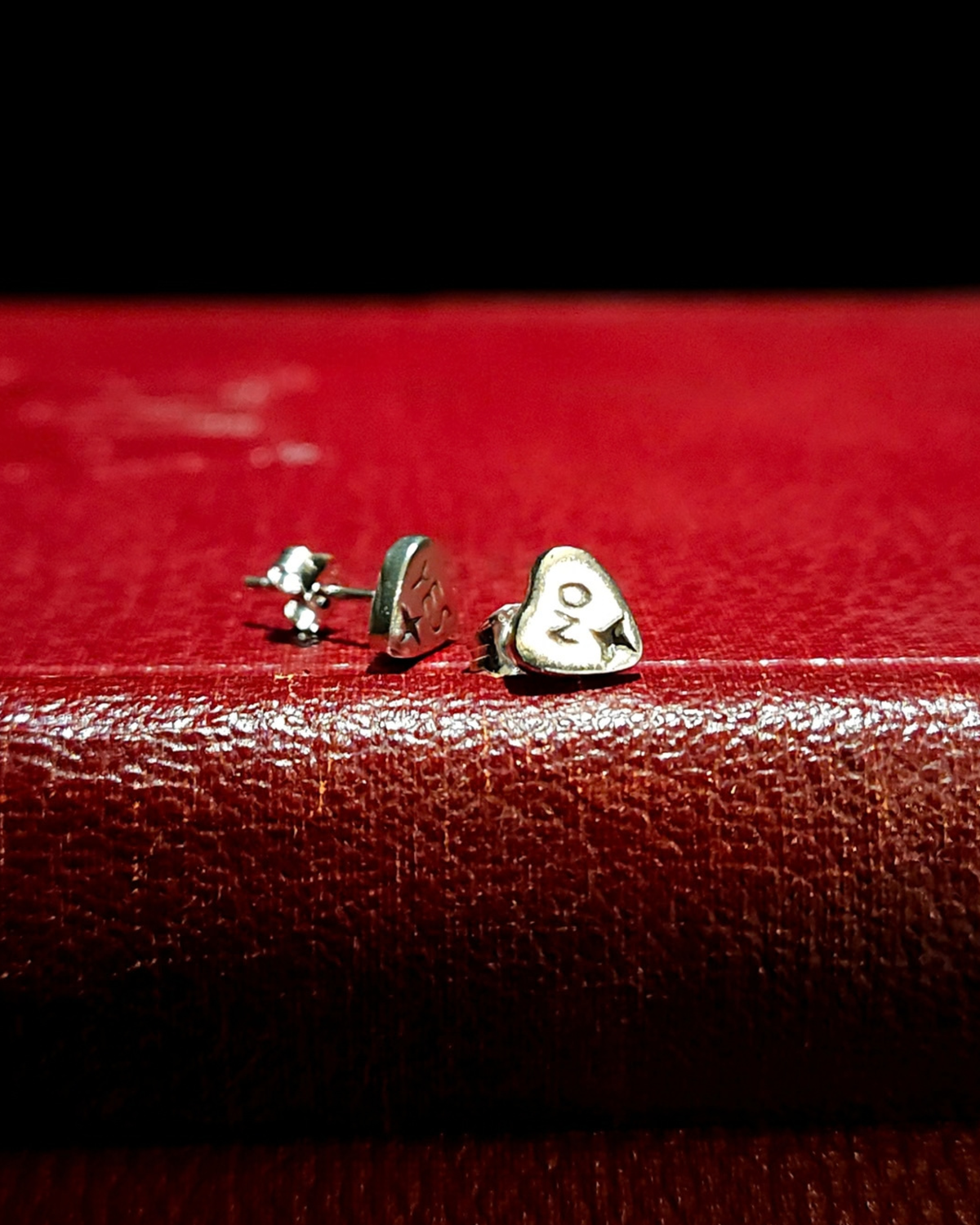 A pair of small, .925 silver spirit board planchette stud earrings sit on a vintage red book with a black background. The earring on the left says "YES" and the one on the right says "NO" and they both have a 4-pointed star at the bottom of each pointer.