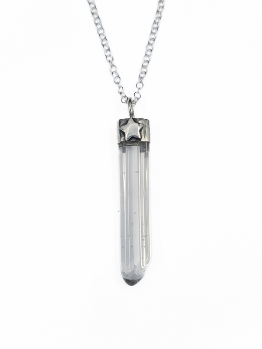 A silver pendant on a chain is displayed on a white background.
It is comprised of a long, thin, clear quartz point coming from a handmade silver setting.
It has a 3D silver star embellishment soldered to the front.