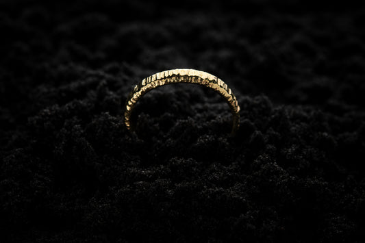 A slim, yellow gold wedding ring sits in black sand to contrast its warm, luscious glow. There are hammered striations around the band like the suns rays. It is half submerged in the sand and breaks through the surface like a golden rainbow.