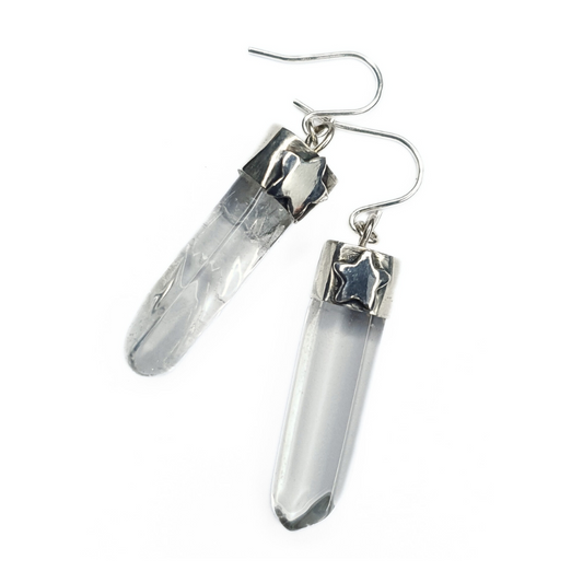 A pair of quartz and silver earrings are displayed on a white background.
They are comprised of a clear quartz point coming from a handmade silver setting with a silver hook.
They each have a 3D silver star embellishment on the front of their silver settings.