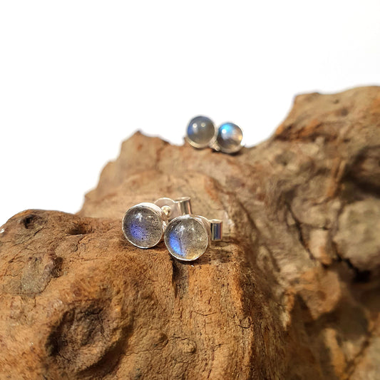 2 pairs of simple, sterling silver and labradorite stud earrings sit on a piece of driftwood. One pair sit in focus, in the foreground and another is out of focus in the background. A gorgeous, bright flash of blue luminescence is visible on both pairs.