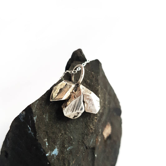 A sterling silver necklace resting on a dark chunk of rock on a white backgorund.. The pendant is made of a cluster of 3 rough-cut nuggets of solid silver. Geometric, unisex, minimalist.