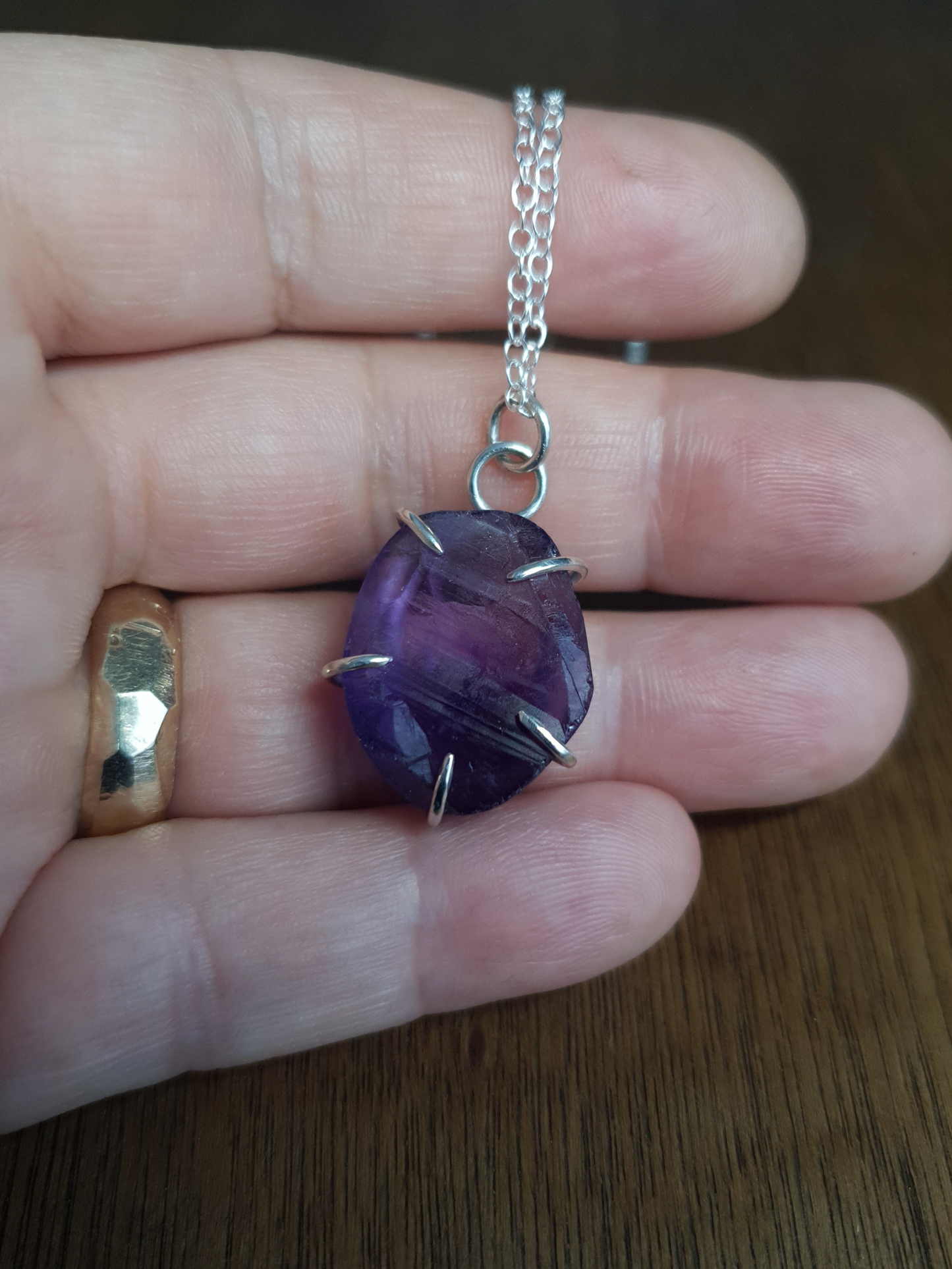 A deep purple amethyst necklace sits on an open hand to demonstrate its size. It has a textured, rough but shiny surface texture and the stone is held by 5 simple, sterling silver claws. It's hanging from a simple sterling silver chain.
