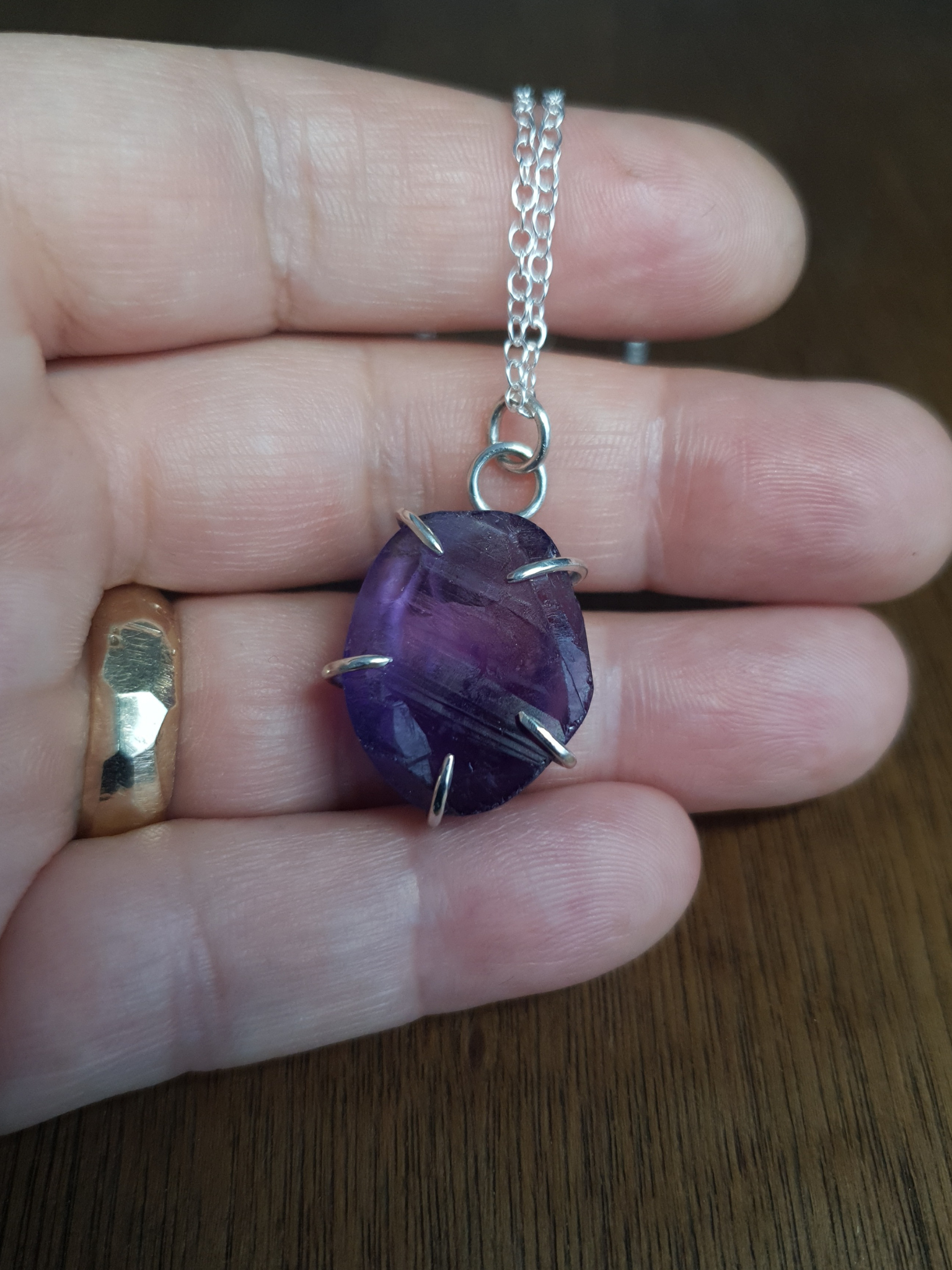 A deep purple amethyst necklace sits on an open hand to demonstrate its size. It has a textured, rough but shiny surface texture and the stone is held by 5 simple, sterling silver claws. It's hanging from a simple sterling silver chain.