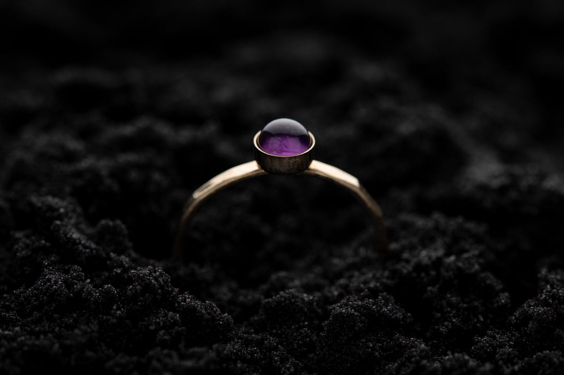 A single amethyst stacking ring with yellow gold band and around amethyst stone, rests gently in soft black sand..