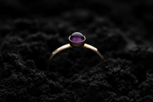 A single amethyst stacking ring with yellow gold band and around amethyst stone, rests gently in soft black sand..