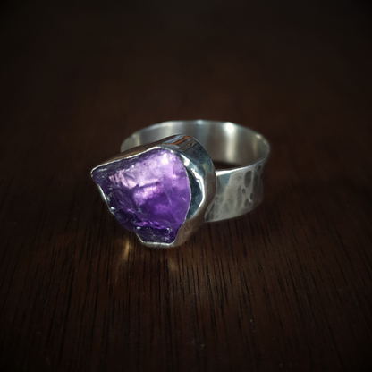 A rough-cut amethyst ring set in a textured sterling silver band, displayed on a warmly toned wooden background. The light illuminates the translucent stone as if by magic