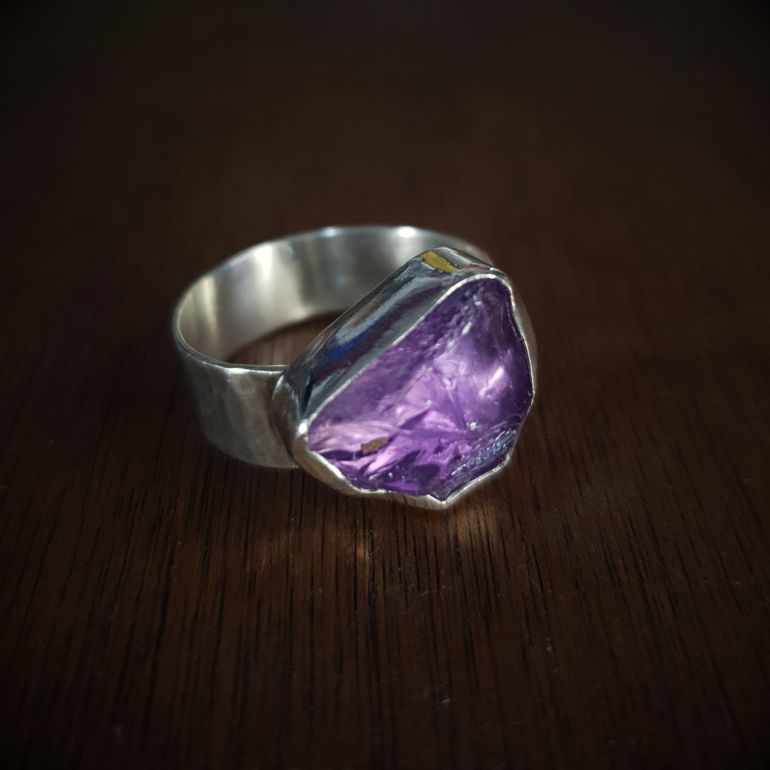 A rough-cut amethyst ring set in a textured sterling silver band, displayed against a wooden background.