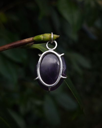 An oval shaped, rough-cut amethyst pendant hangs from a thin budded twig, against a dark green natural background. It is a rich purple colour and is photographed from the back to show how its simple design allows the light to enter the stone and enhance its deep colours and natural textures. The setting is made from recycled sterling silver.