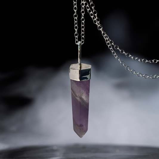 A sterling silver chain necklace featuring a polished purple amethyst crystal point pendant with a silver cap, hanging against a dark, smoky background.
Wilde Folk Jewellery is inspired by nature, folklore & mysticism. A queer-owned brand using responsibly sourced gemstones & recycled precious metals.