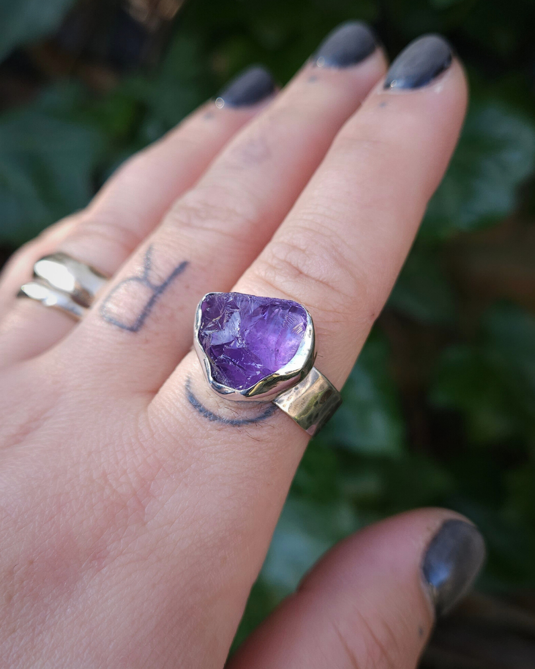 A hand wears a rough cut amethyst ring on the forefinger against a dark green natural, botanical background.