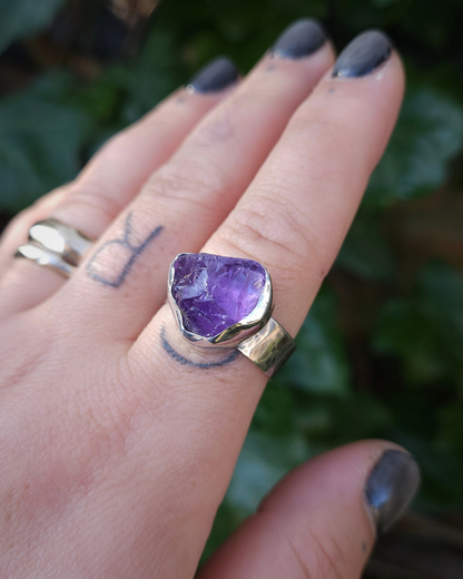 A hand wears a rough cut amethyst ring on the forefinger against a dark green natural, botanical background.