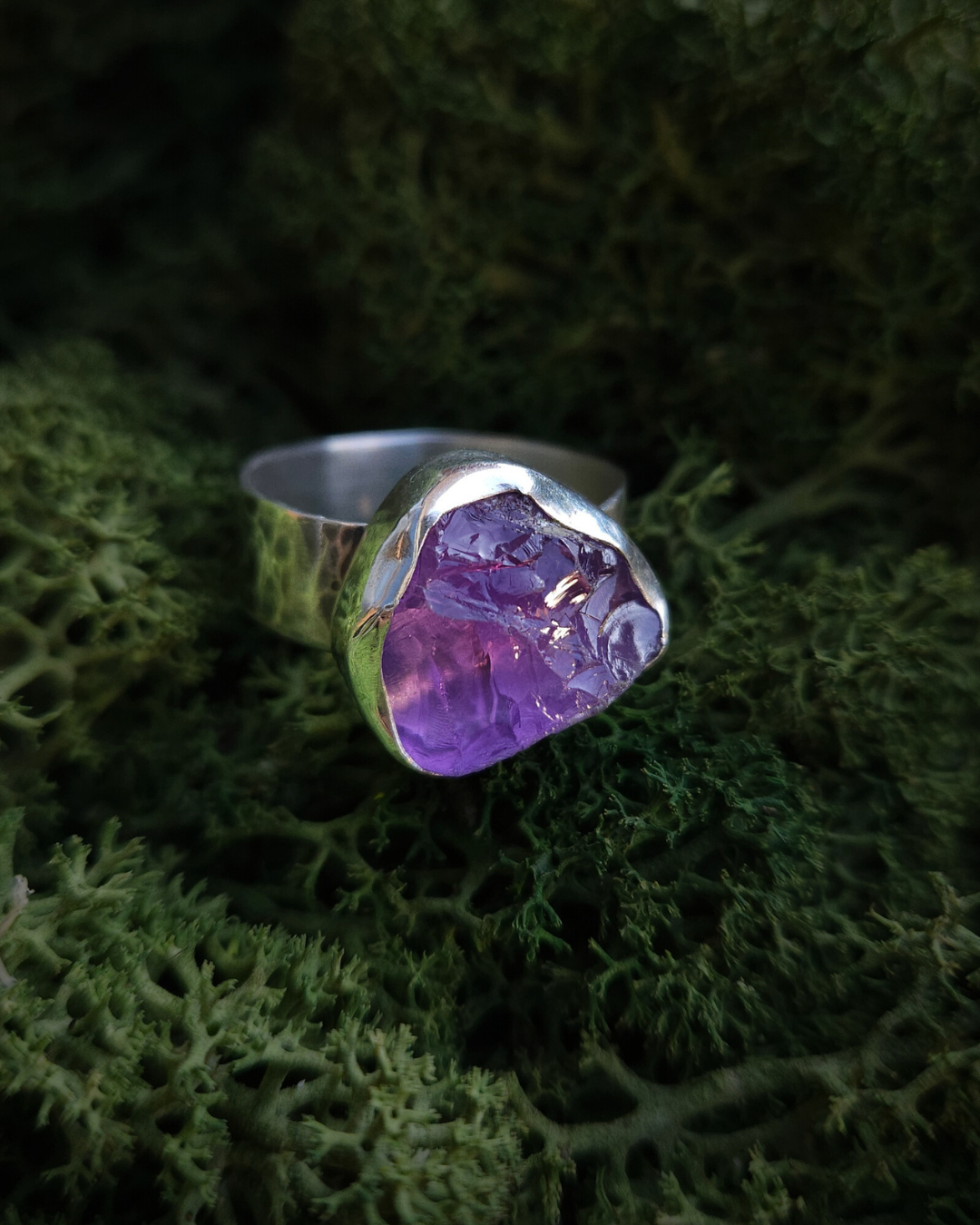 A rough-cut amethyst and sterling silver ring sits on a soft bed of vibrant-green moss and is illuminated by sunlight.