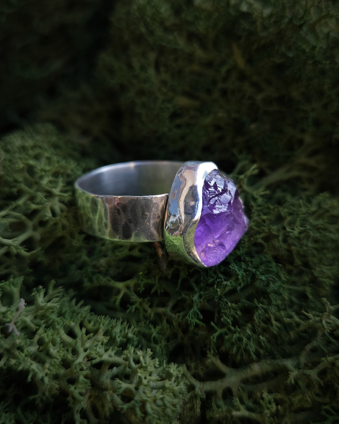 A rough-cut amethyst and sterling silver ring sits on a soft bed of vibrant-green moss and is illuminated by sunlight. The band has hammered, oxidised texture and is made from recycled sterling silver.