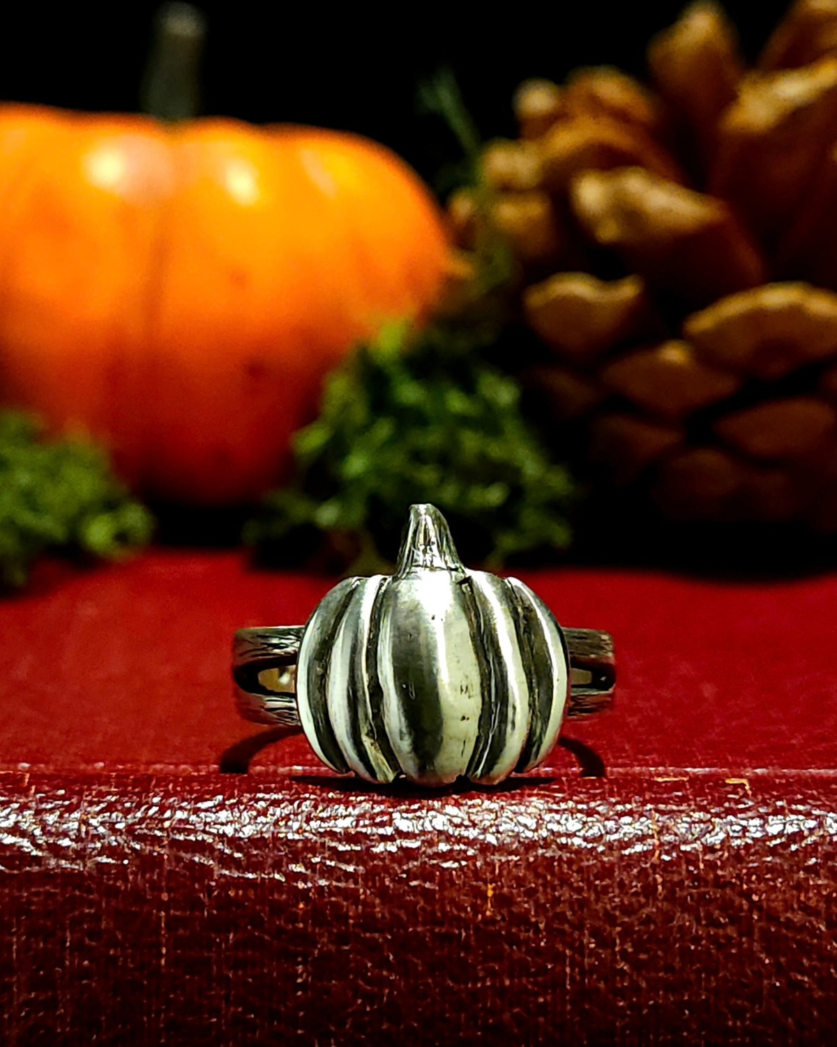 A sterling silver autumn pumpkin ring with dark oxidised detailing, displayed on a textured vintage red book with a moss, an orange pumpkin and pine cone in the background out of focus.