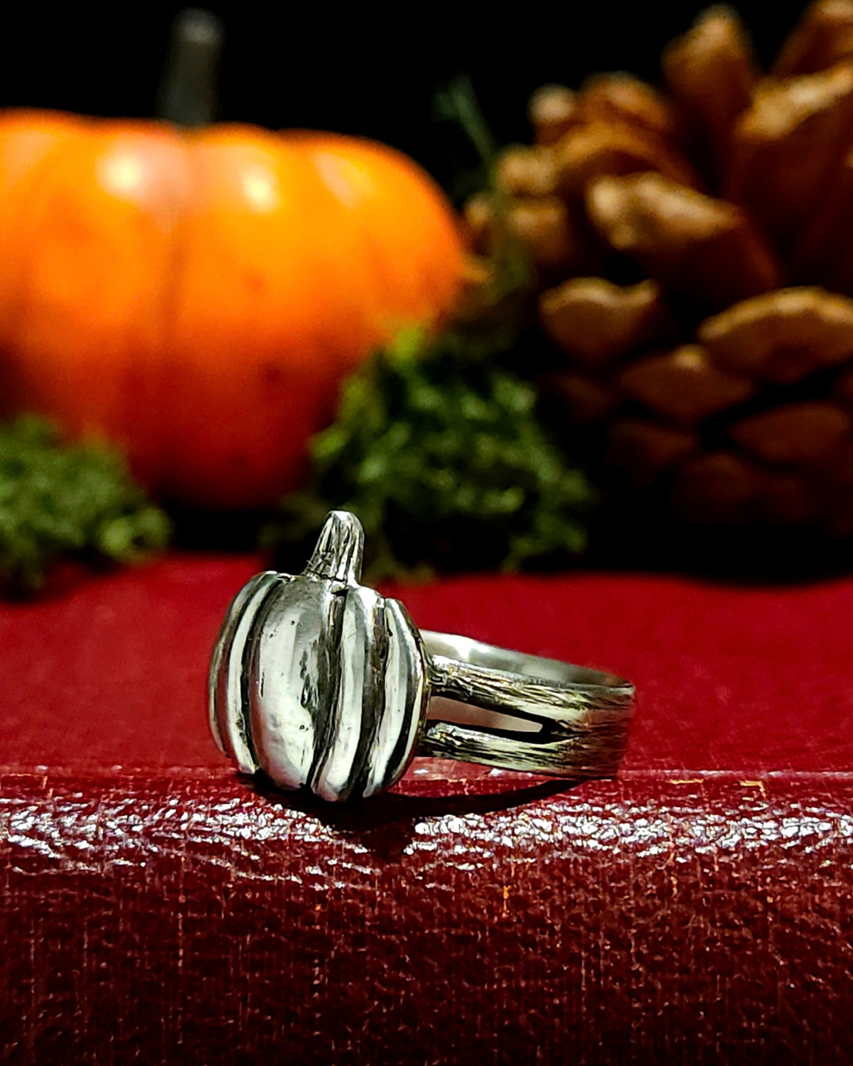 A .925 silver halloween pumpkin ring with dark oxidised detailing, displayed on a textured vintage red book. The band and pumpkin have been carved to have a natural, textured surface. There is moss, an orange pumpkin and pine cone in the background out of focus to create an autumnal atmosphere.