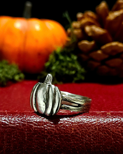 A .925 silver halloween pumpkin ring with dark oxidised detailing, displayed on a textured vintage red book. The band and pumpkin have been carved to have a natural, textured surface. There is moss, an orange pumpkin and pine cone in the background out of focus to create an autumnal atmosphere.