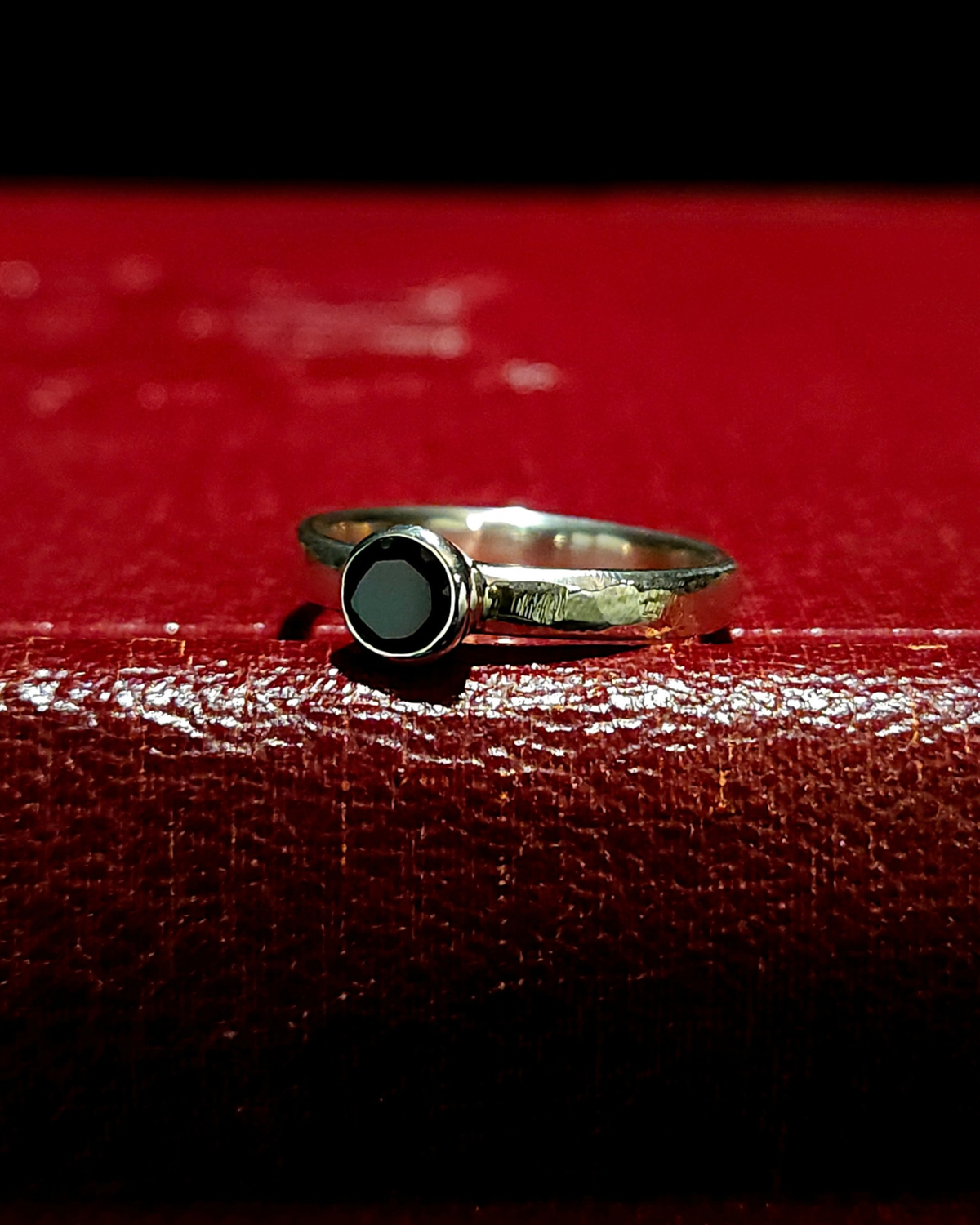A gothic , .925 silver solitaire ring with a stylistic rough-filed band and faceted black spinel lays on its side on a dark red, vintage book.