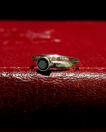 A gothic , .925 silver solitaire ring with a stylistic rough-filed band and faceted black spinel lays on its side on a dark red, vintage book.