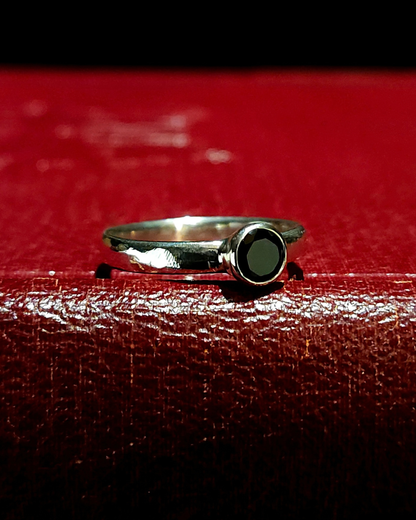 A sterling silver solitaire ring with a stylistic rough-filed band and faceted black spinel lays on its side on a dark red, vintage book.