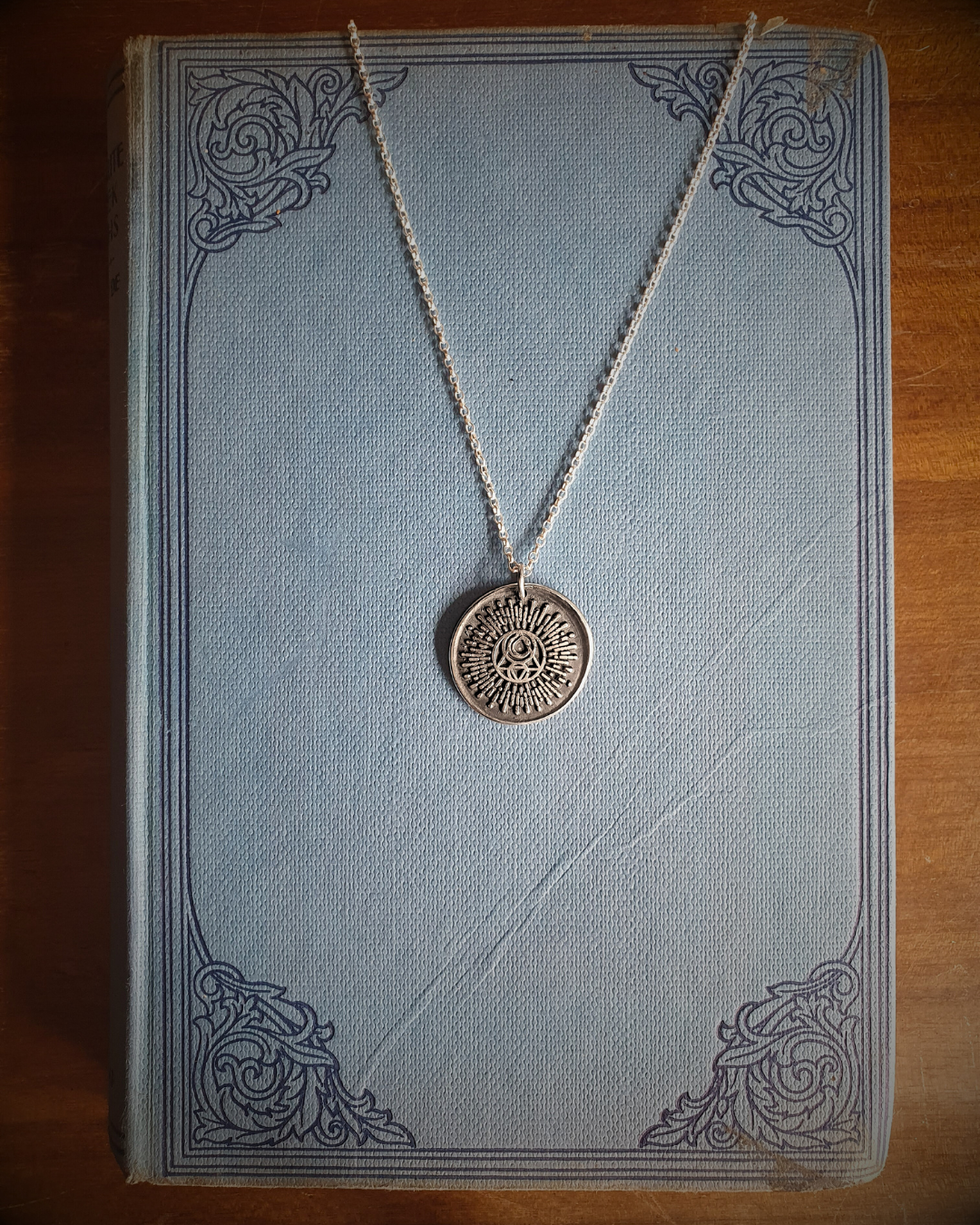 A silver wax seal pendant featuring the Campfire & Compass Sigil logo with a sun and moon and alchemical symbols for fire and water, displayed on a vintage blue book.
A silver pendant with a circular design featuring solar and lunar elements, displayed on a stack of vintage books.