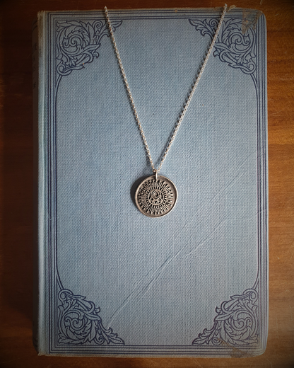 A silver wax seal pendant featuring the Campfire & Compass Sigil logo with a sun and moon and alchemical symbols for fire and water, displayed on a vintage blue book.
A silver pendant with a circular design featuring solar and lunar elements, displayed on a stack of vintage books.