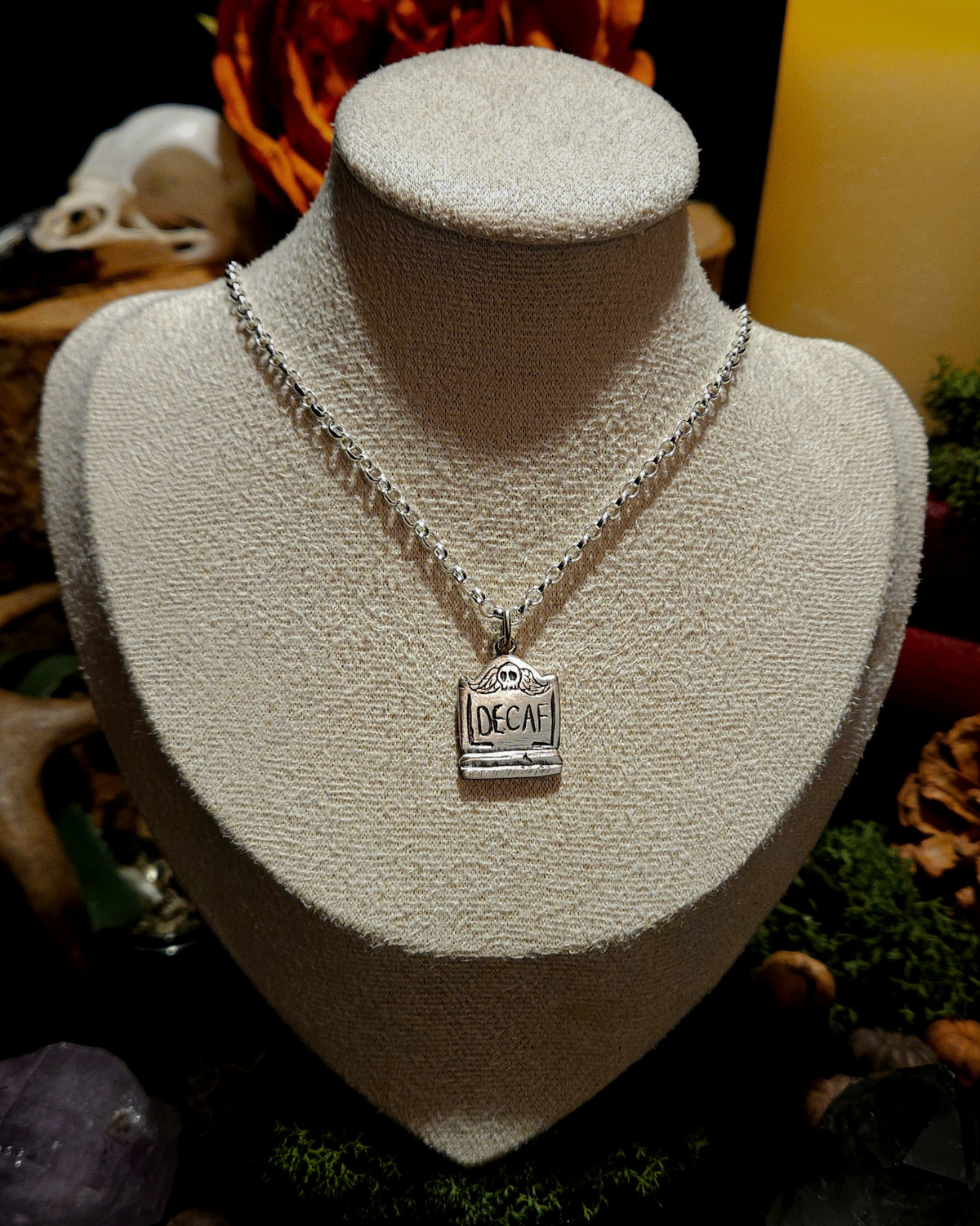 A .925 silver tombstone pendant featuring a skull, and engraved with 'DECAF', is displayed on a beige vintage style bust. The display is set against a dark, warm & autumnal background with candles, natural elements, moss, crystals and other macabre and dark academia themed items