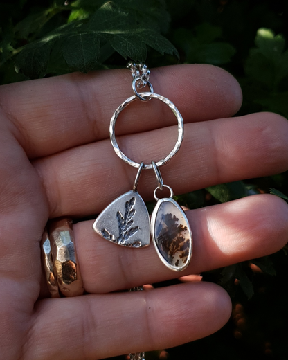 Silver necklace with a solid silver triangle, imprinted with a cypress sprig, and dendritic agate stone pendant hang from a silver circlet and sit in an open palm to demonstrate size. The dendritic agate is translucent with dark brown dendrites. The hand is wearing two gold rings and the background is comprised of natural greenery.