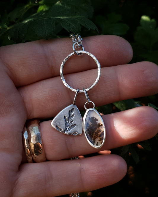 Silver necklace with a solid silver triangle, imprinted with a cypress sprig, and dendritic agate stone pendant hang from a silver circlet and sit in an open palm to demonstrate size. The dendritic agate is translucent with dark brown dendrites. The hand is wearing two gold rings and the background is comprised of natural greenery.