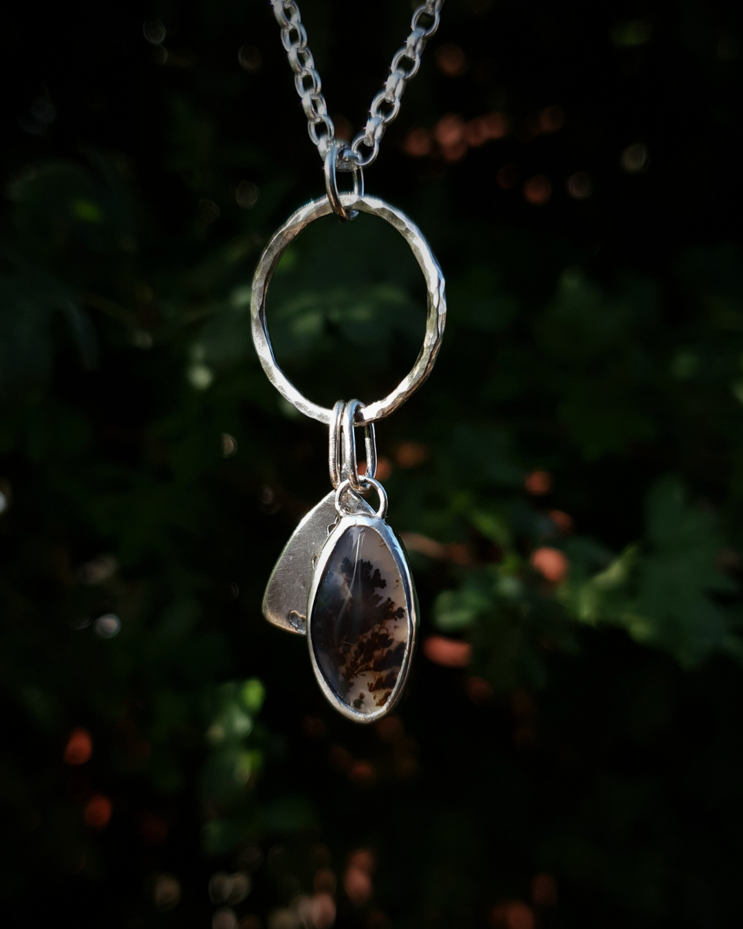 Silver pendant comprised of a silver circlet from which a solid silver triangle, imprinted with a cypress sprig, and dendritic agate stone pendant hang from a silver chain. The dendritic agate is translucent with dark brown dendrites. The background is out of focus natural greenery.