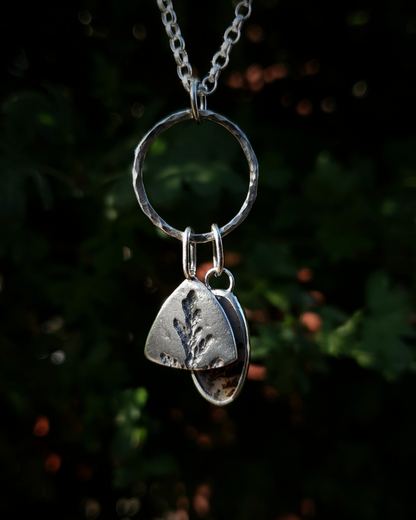 Silver pendant comprised of a silver circlet from which a solid silver triangle, imprinted with a cypress sprig, and dendritic agate stone pendant hang from a silver chain. The dendritic agate is translucent with dark brown dendrites. The background is out of focus natural greenery.