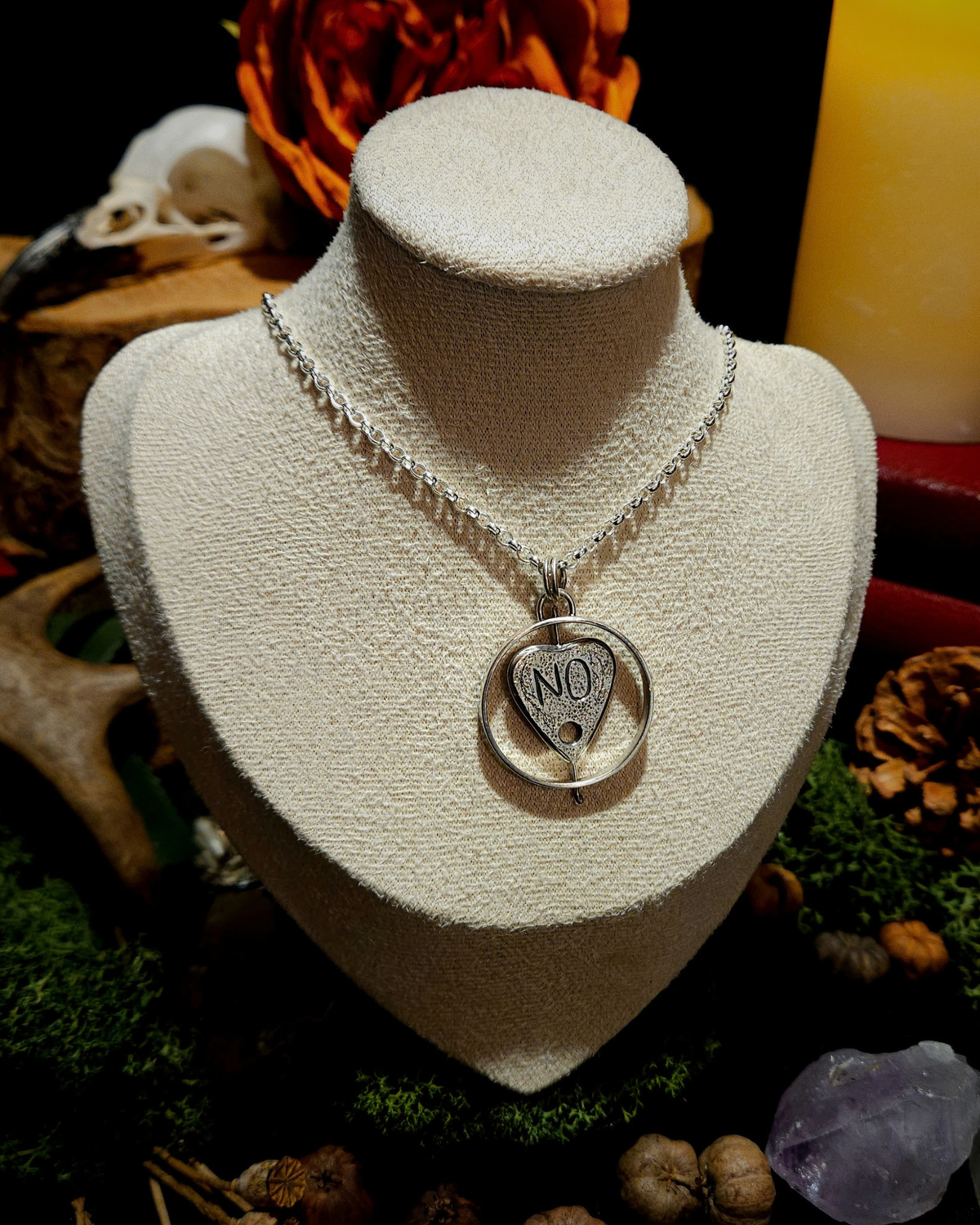 A large sterling silver ouija planchette necklace is displayed on a beige velvet bust amongst occult, mystical and nature based autumnal decor. The planchette is surrounded by a thick circle, that it can spin within. This side of the planchette reads "NO".