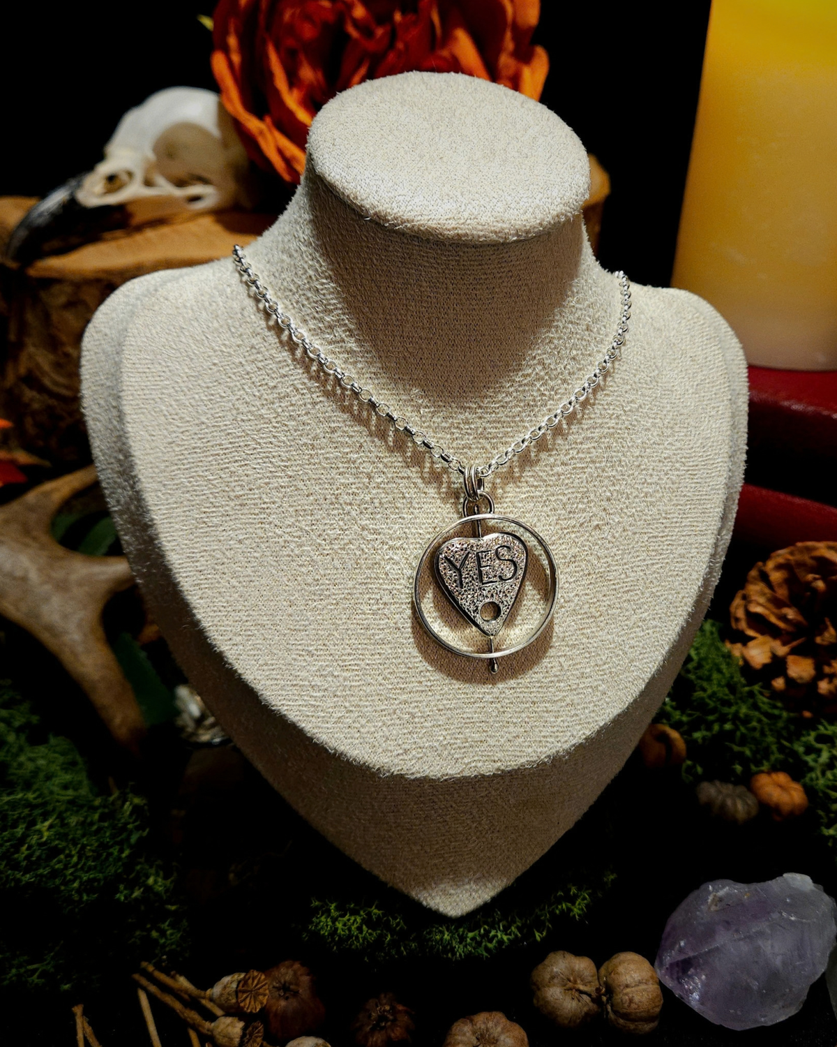 A large ouija planchette necklace is displayed on a cream velvet bust amongst witchy, mystical and nature based autumnal decor. The planchette is surrounded by a thick circle, that it can spin within. This side of the planchette reads "YES".